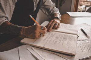 Close-up of a tattooed hand writing in a notebook with pencil and papers around.