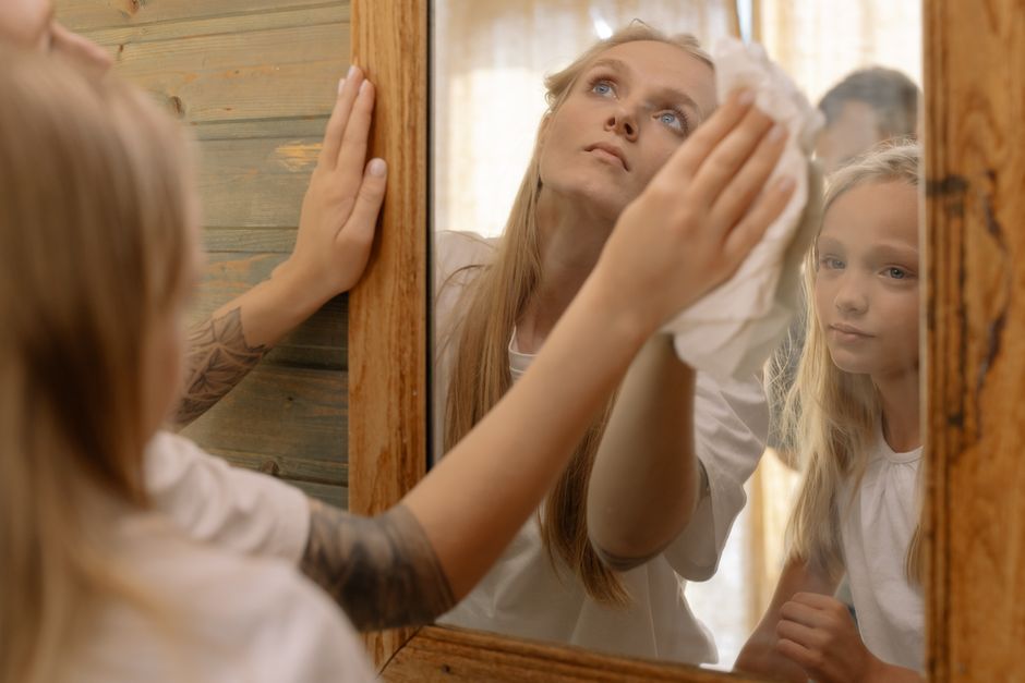 A mother wiping a mirror while her daughter watches, reflecting family bonding and care.