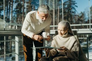 Senior couple enjoying coffee and reading in a sunlit room with large windows.