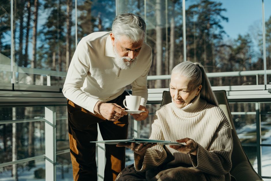 Senior couple enjoying coffee and reading in a sunlit room with large windows.
